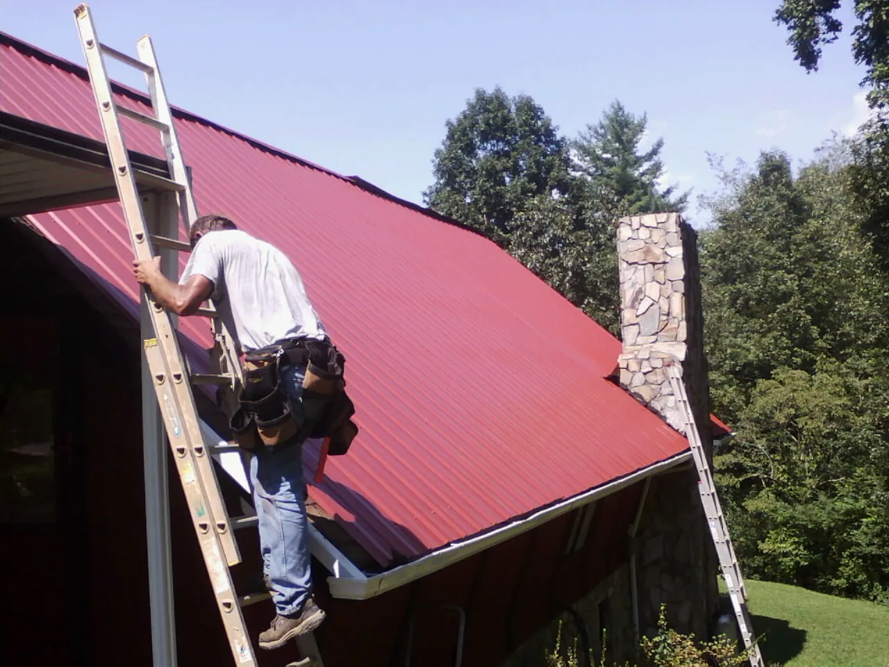 Roofer on a ladder beside a red metal roof and stone chimney — Soffit & Fascia Repair by Roofing Brothers in Conneaut Lake